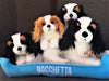 Four Cavalier King Charles Spaniel plush toys in black‑tan and white‑brown colors sitting on blue Bocchetta dog bed