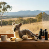 Plush kangaroo toy with binoculars and a mug on a windowsill with a scenic view.