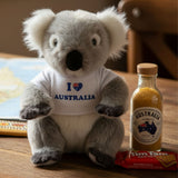 Plush koala wearing a 'I ❤️ Australia' shirt with a bottle of Australian gold and Tim Tam on a wooden surface.