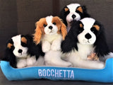 Four Cavalier King Charles Spaniel plush toys in black‑tan and white‑brown colors sitting on blue Bocchetta dog bed