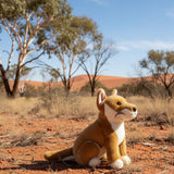 Plush toy dingo in a desert landscape with trees and blue sky.